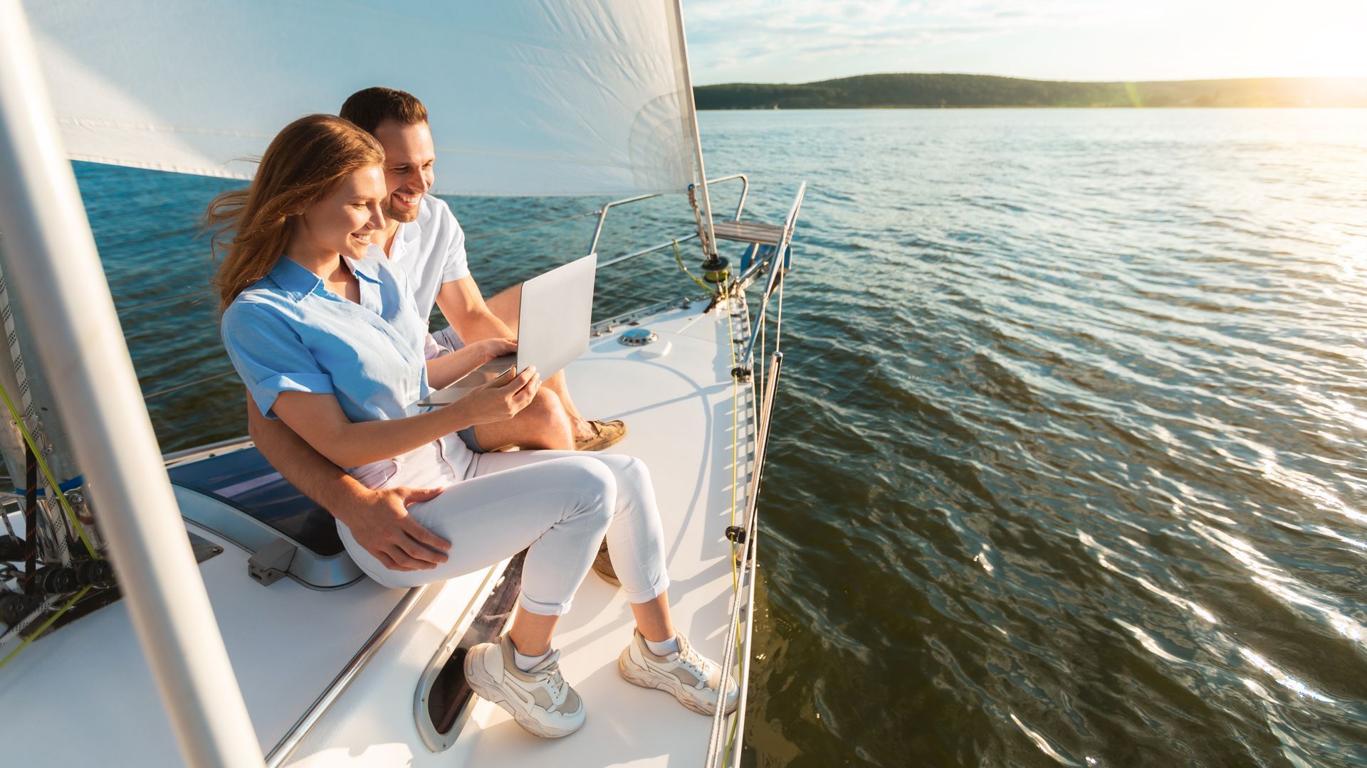 A man and a woman are sitting on a sailboat looking at a laptop.