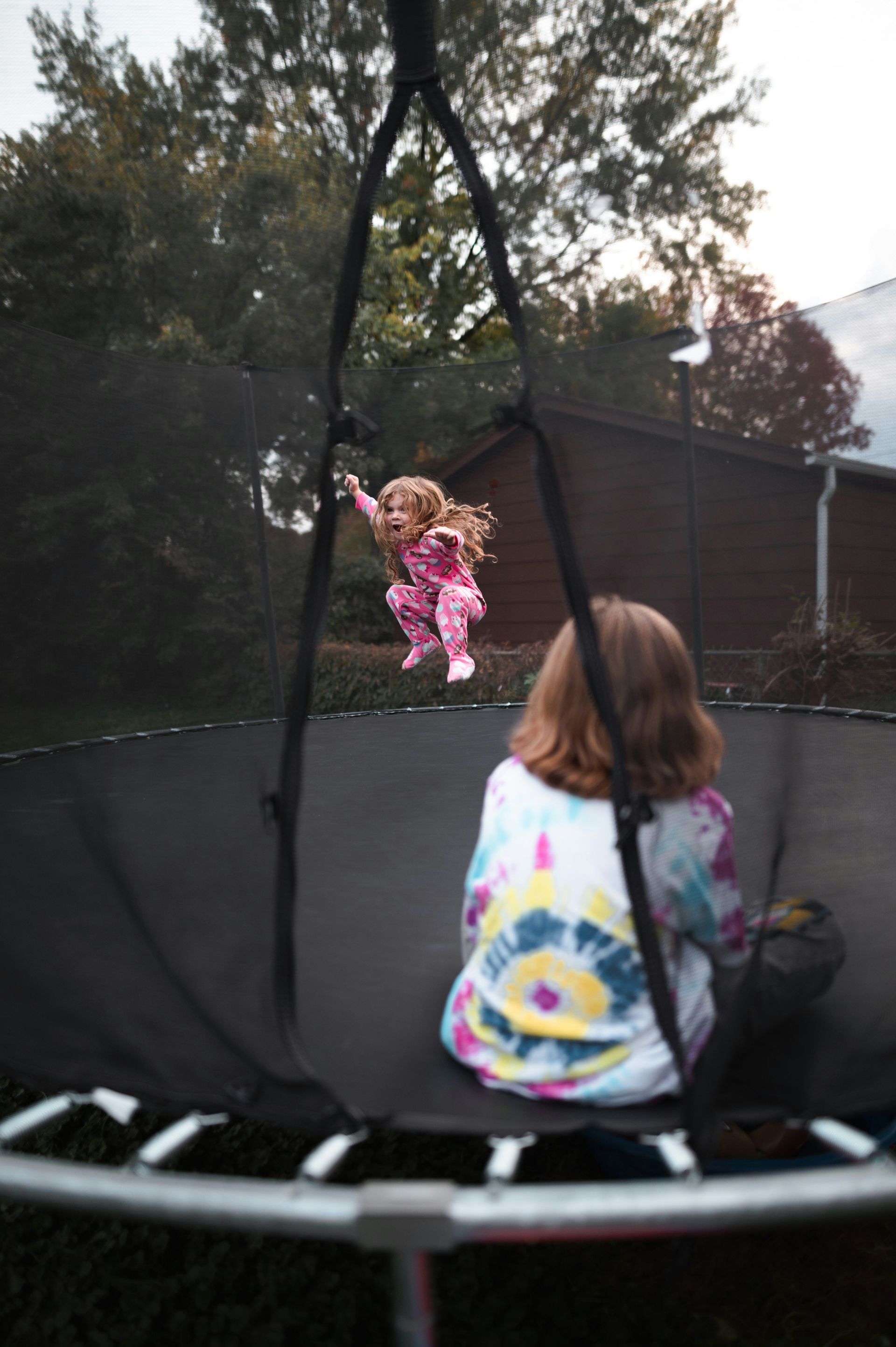 A little girl is jumping on a trampoline while another little girl watches.