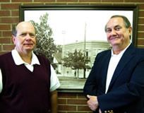 Two men are standing next to each other in front of a picture on a brick wall.