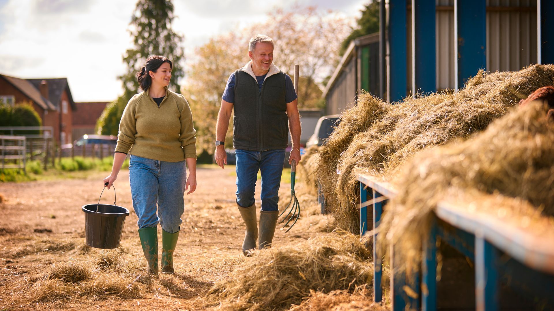 A man and a woman are walking in a field next to a pile of hay.