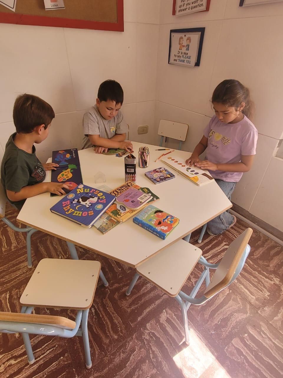 Tres niños están sentados alrededor de una mesa hexagonal en una habitación, entretenidos con libros y rompecabezas.