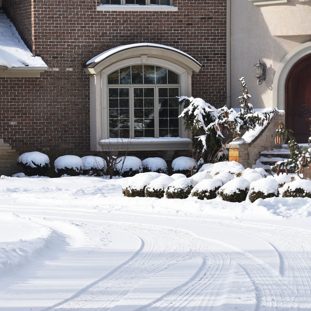 A snowy driveway in front of a brick house with snow covered shrubs