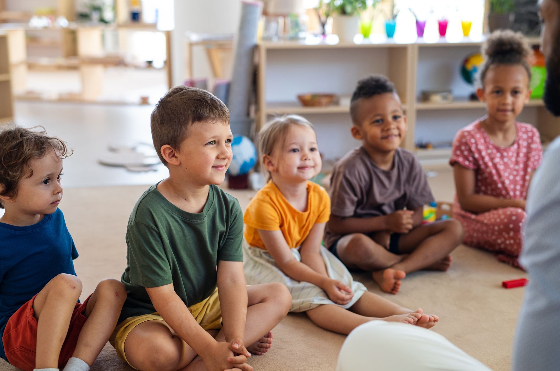 A group of children are sitting on the floor in a circle listening to a teacher.