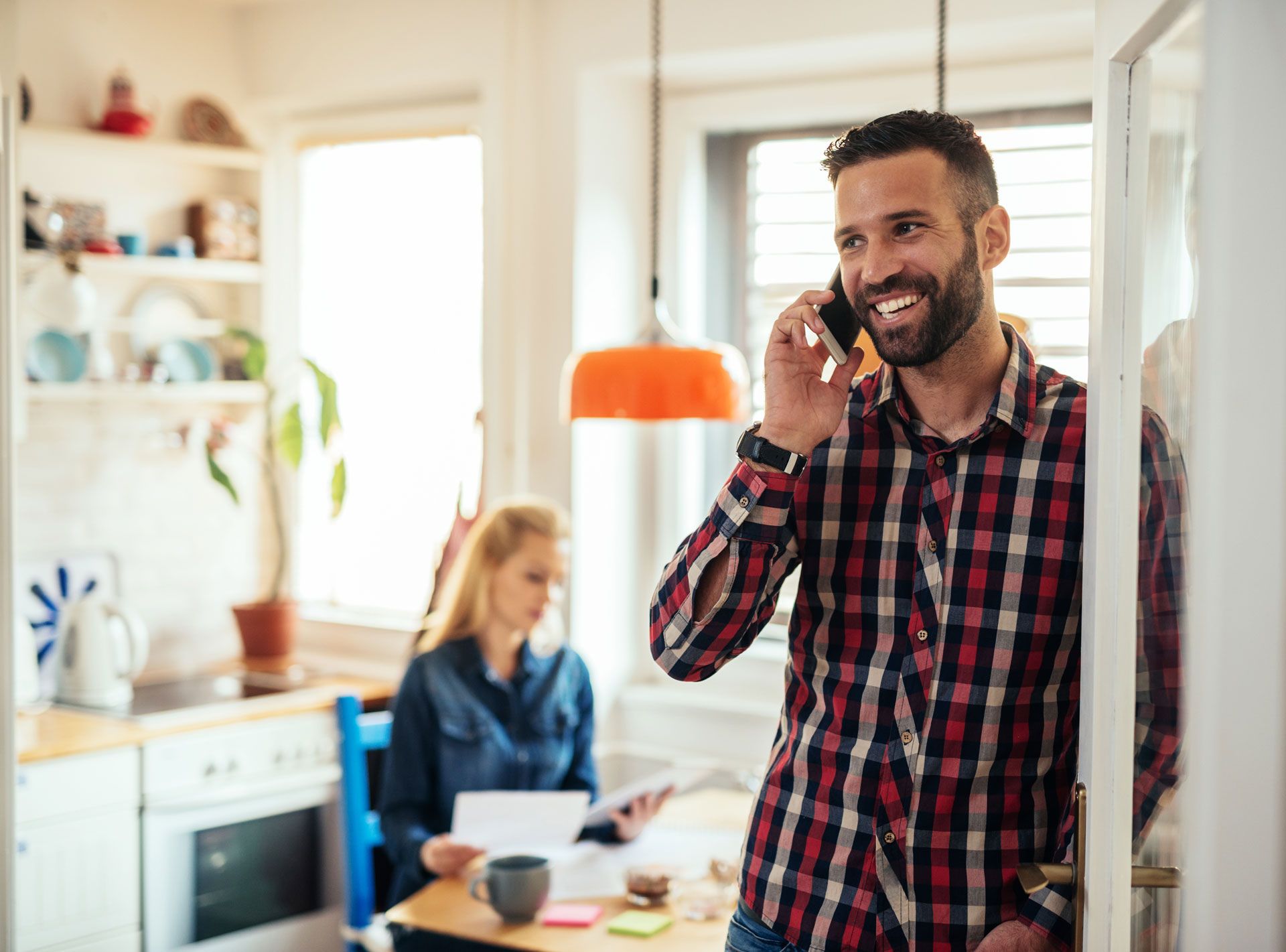 A man is talking on a cell phone in a kitchen while a woman sits at a table.