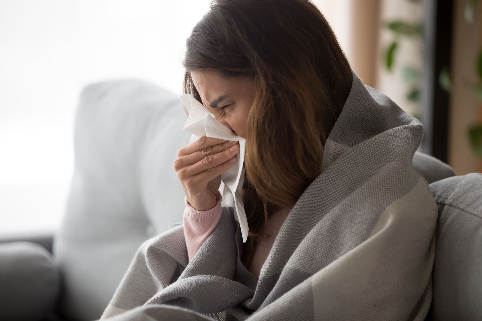 A woman is sitting on a couch blowing her nose into a napkin.