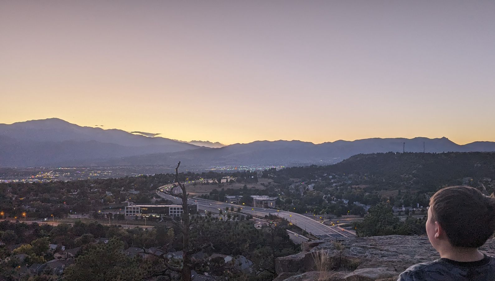 A man is standing on top of a hill overlooking a city at sunset.