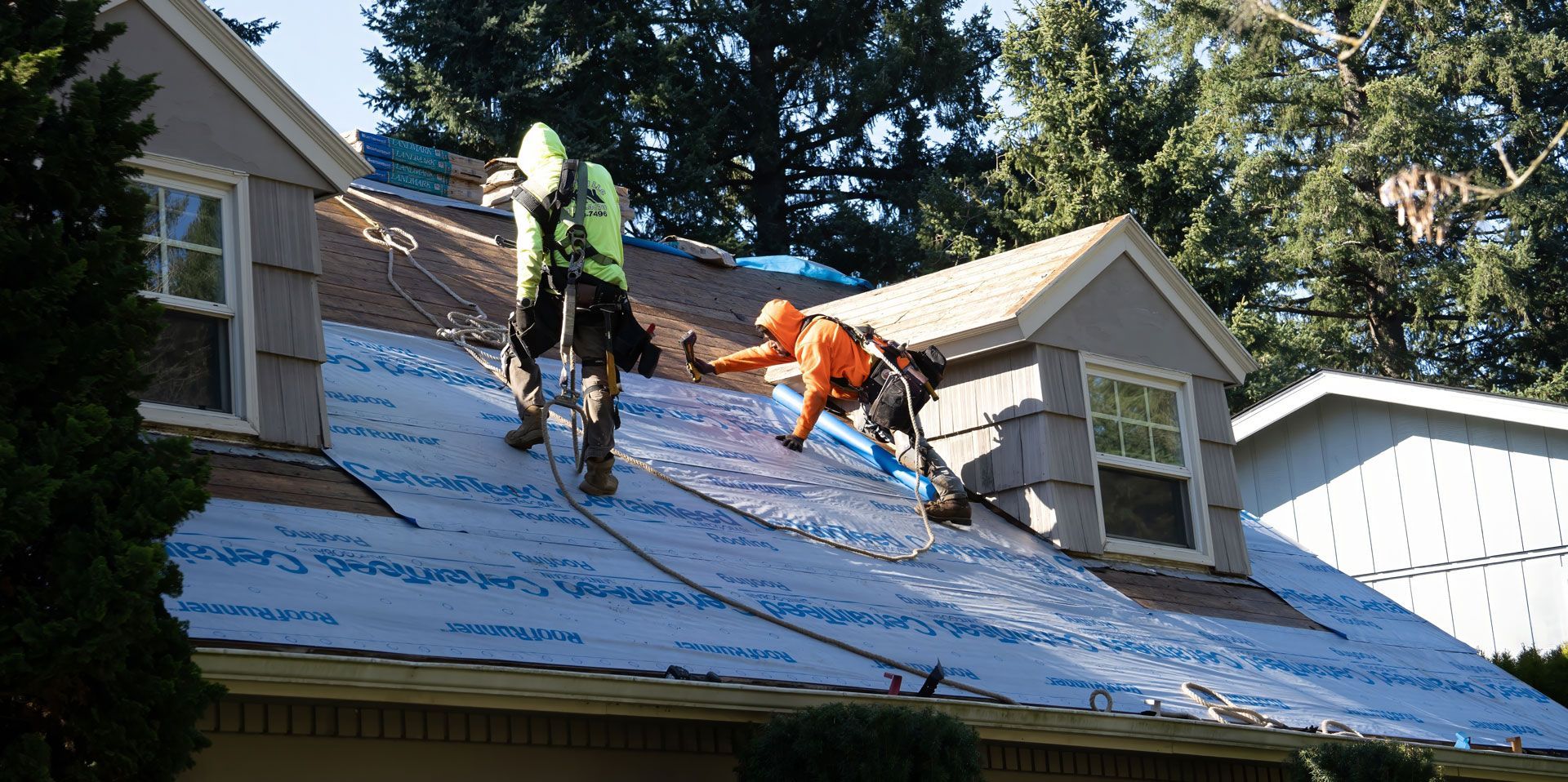 Two roofers in safety gear working on a house roof, installing roofing paper.