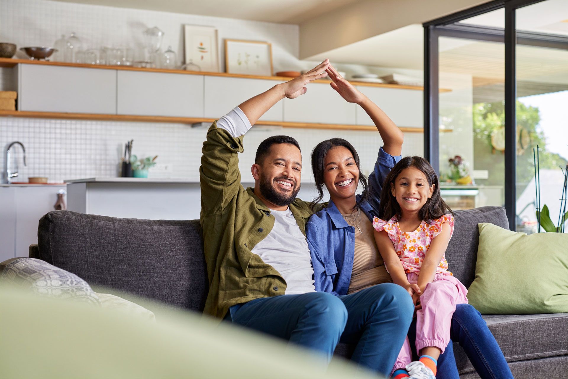 Family of three smiles on a couch. Parents make a house shape above the daughter's head in a bright living room.