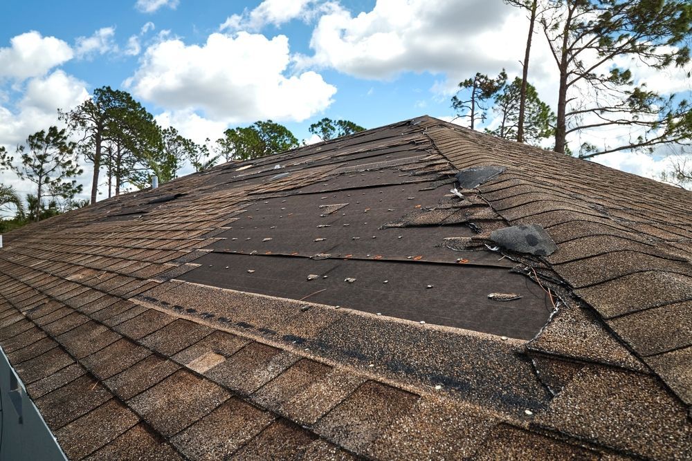 A roof with shingles that are missing and trees in the background