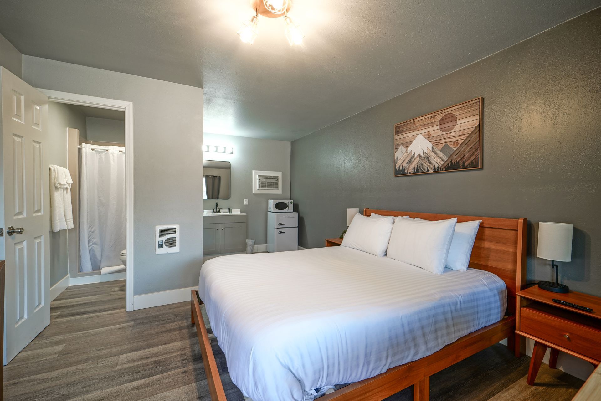 Bedroom with a bed, small table, microwave, and television. Grey walls, light wood floor, and soft lighting.