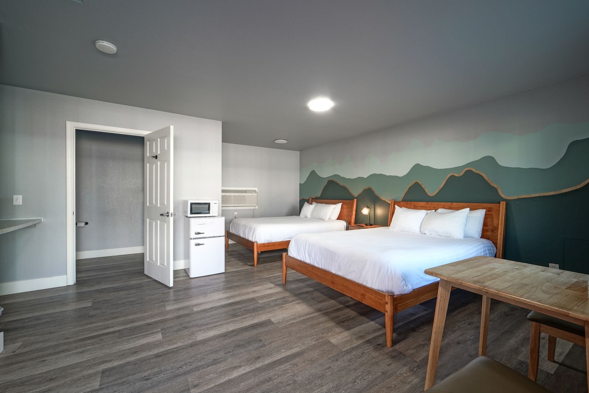 Bedroom with two beds, mountain mural, and wooden table. White bedding, gray walls, and light wood floor.