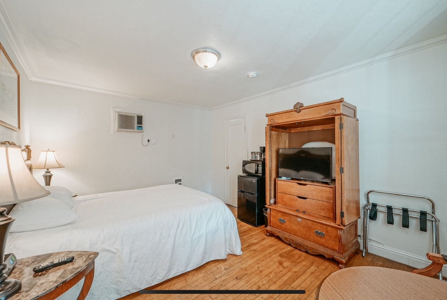 Hotel room with bed, wooden cabinet with TV, mini-fridge, luggage rack. White walls, wood floor.