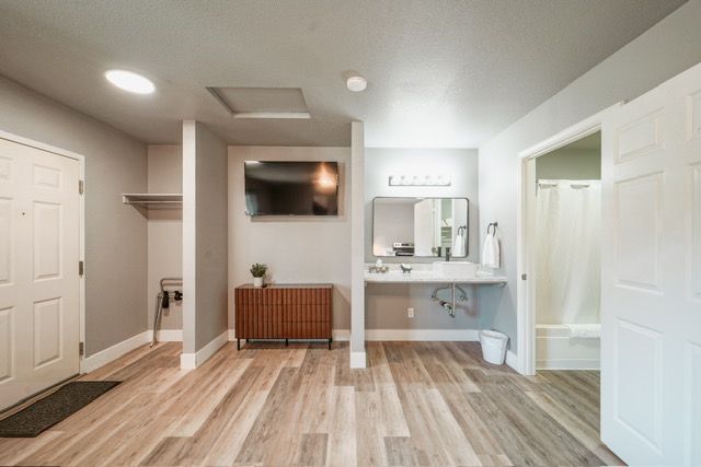 Modern entryway with light wood floors, wall-mounted TV, white closet doors, and a small desk with mirror.