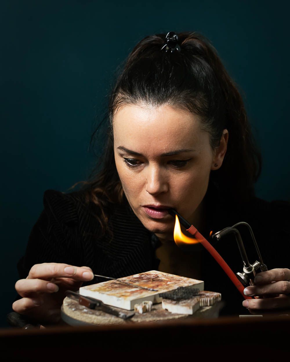 Vrouw soldeert metaal, houdt een brander vast, geconcentreerde uitdrukking, donkere omgeving.