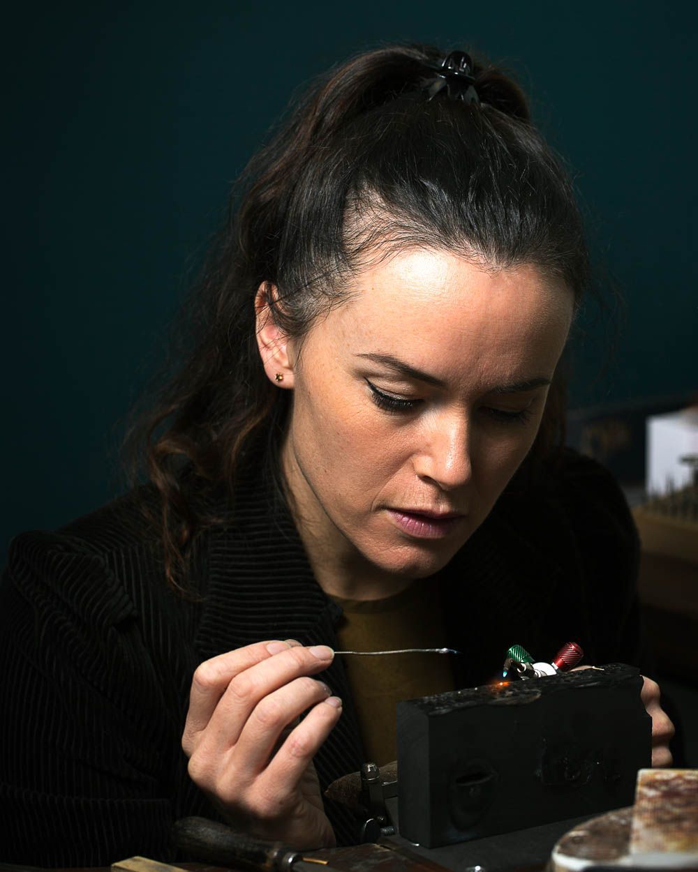 Vrouw werkt geconcentreerd aan sieraden, houdt gereedschap vast, donkere achtergrond.