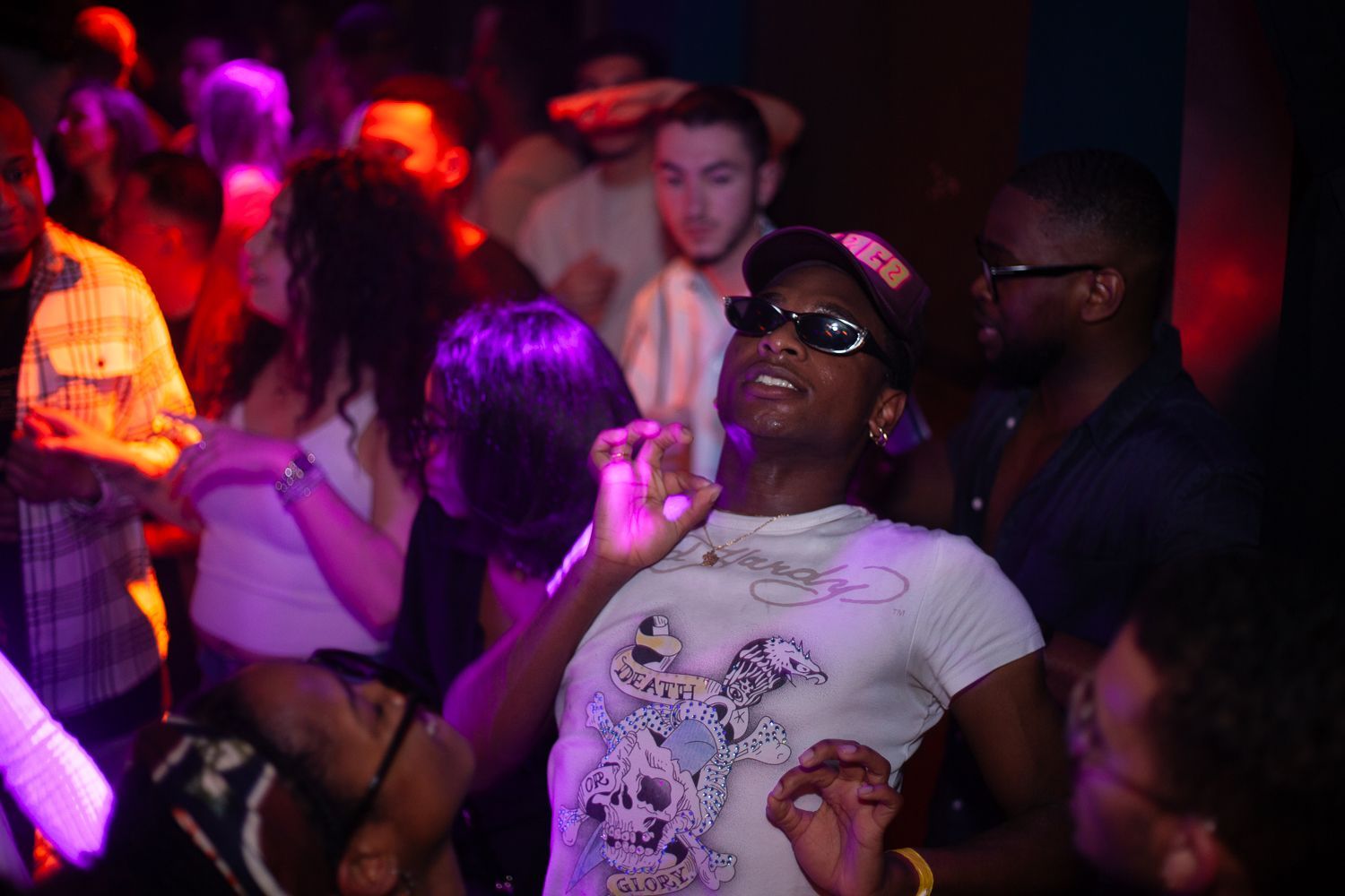 Mensen dansen in een drukke nachtclub met kleurrijke lichten. Een persoon in een wit overhemd en een zonnebril staat centraal, dansend met opgeheven armen.