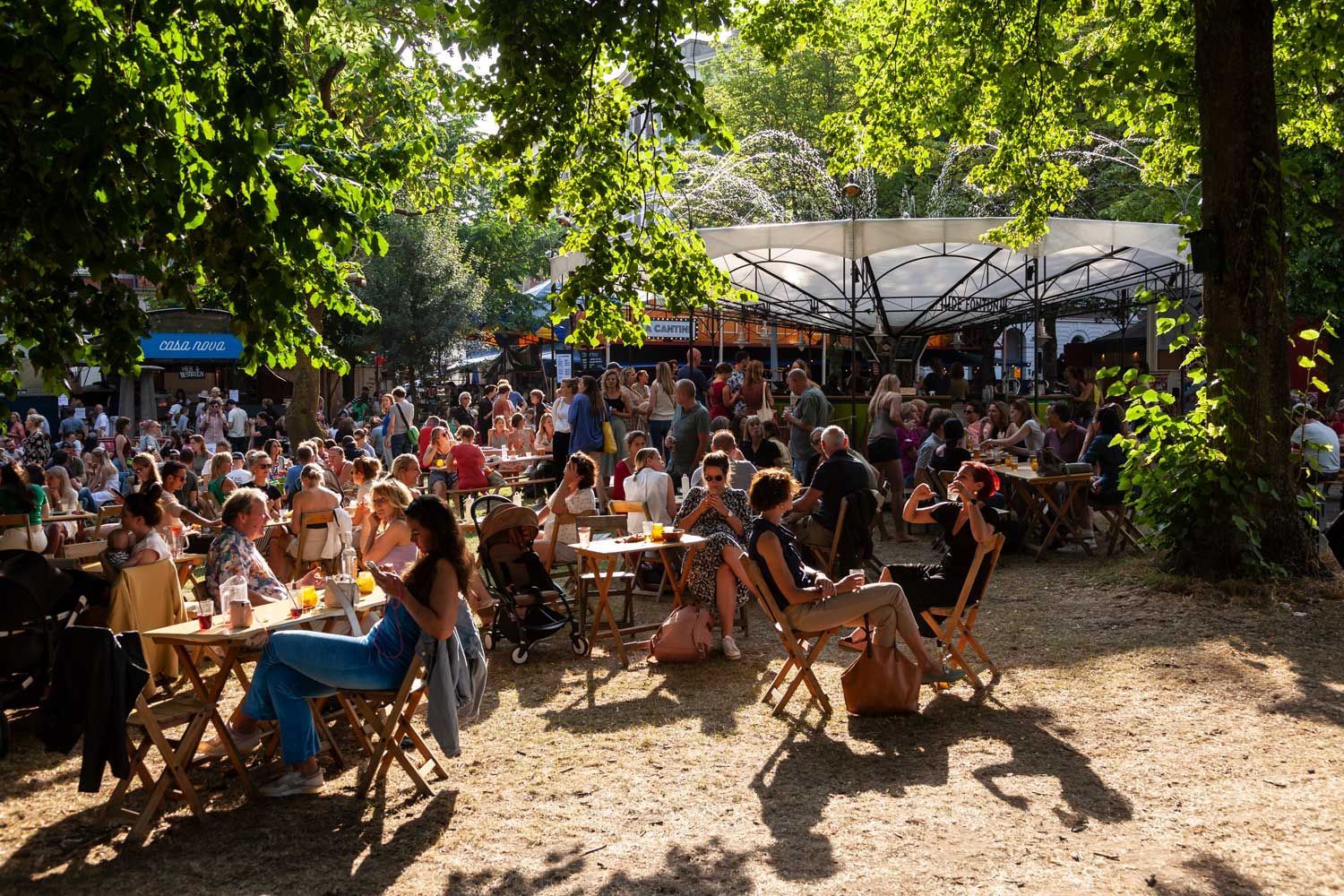 Terras met veel mensen aan tafeltjes onder de bomen. Zonneschijn.
