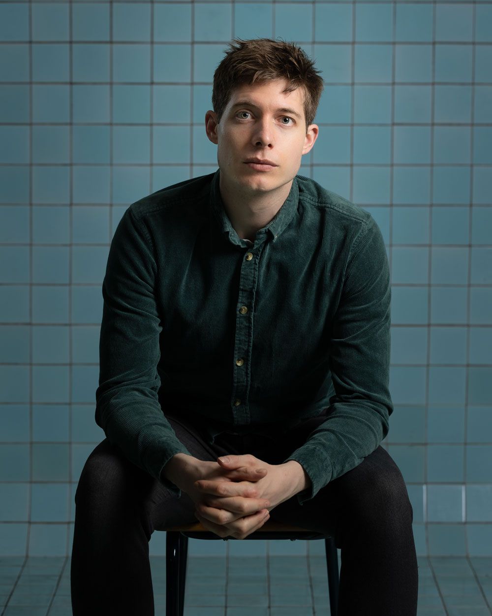 Man in groen shirt zittend op een kruk, handen gevouwen. Blauwe tegelachtergrond.