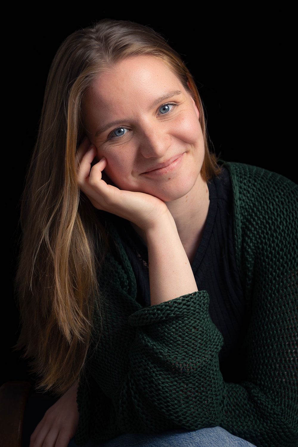 Vrouw met lang haar, haar kin op haar hand rustend, glimlachend. Ze draagt een groene trui, zwarte achtergrond.
