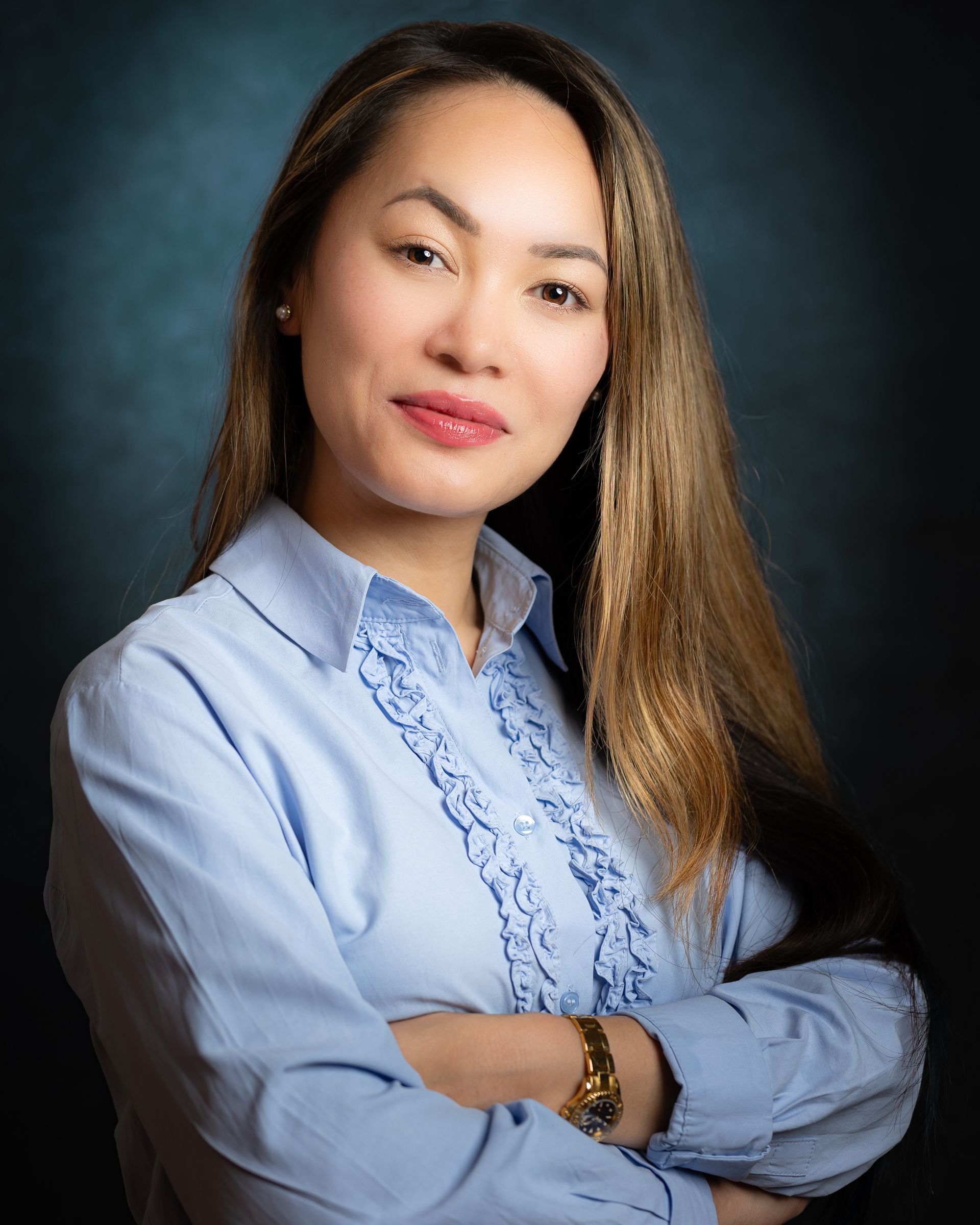 Vrouw met lang bruin haar, lichtblauw shirt, armen over elkaar, lachend, tegen een donkerblauwe achtergrond.