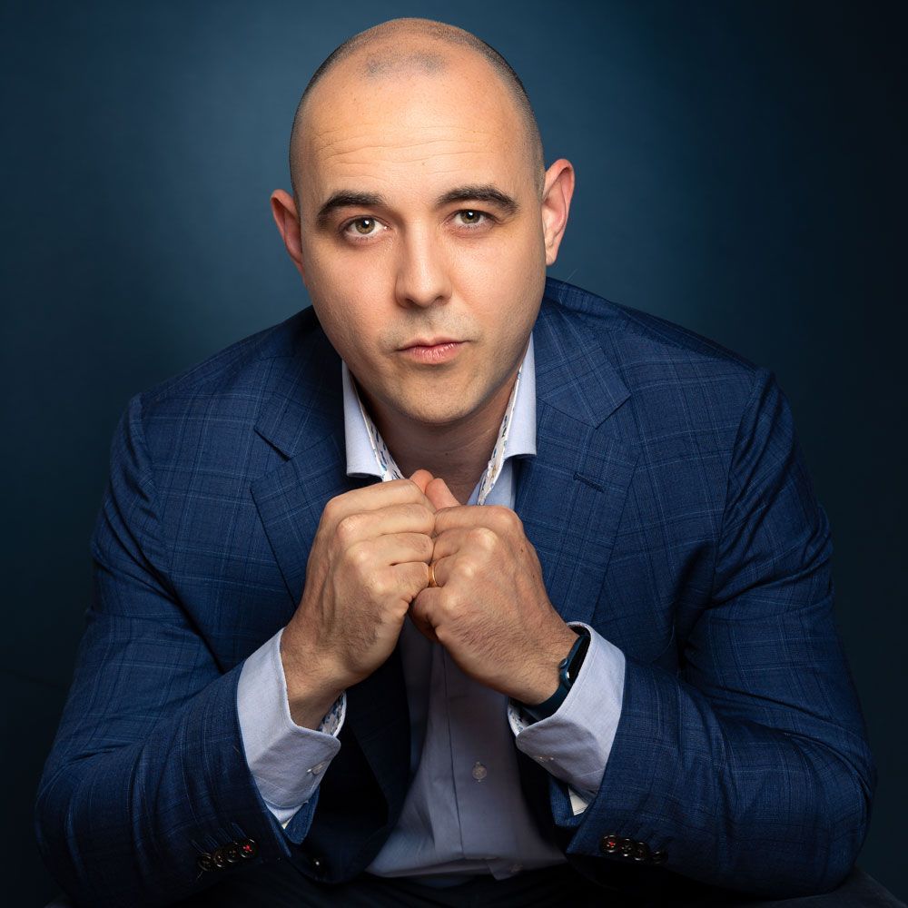 Man in blauwe blazer, handen ineengevouwen, kijkt naar de camera. Tegen een donkerblauwe achtergrond.