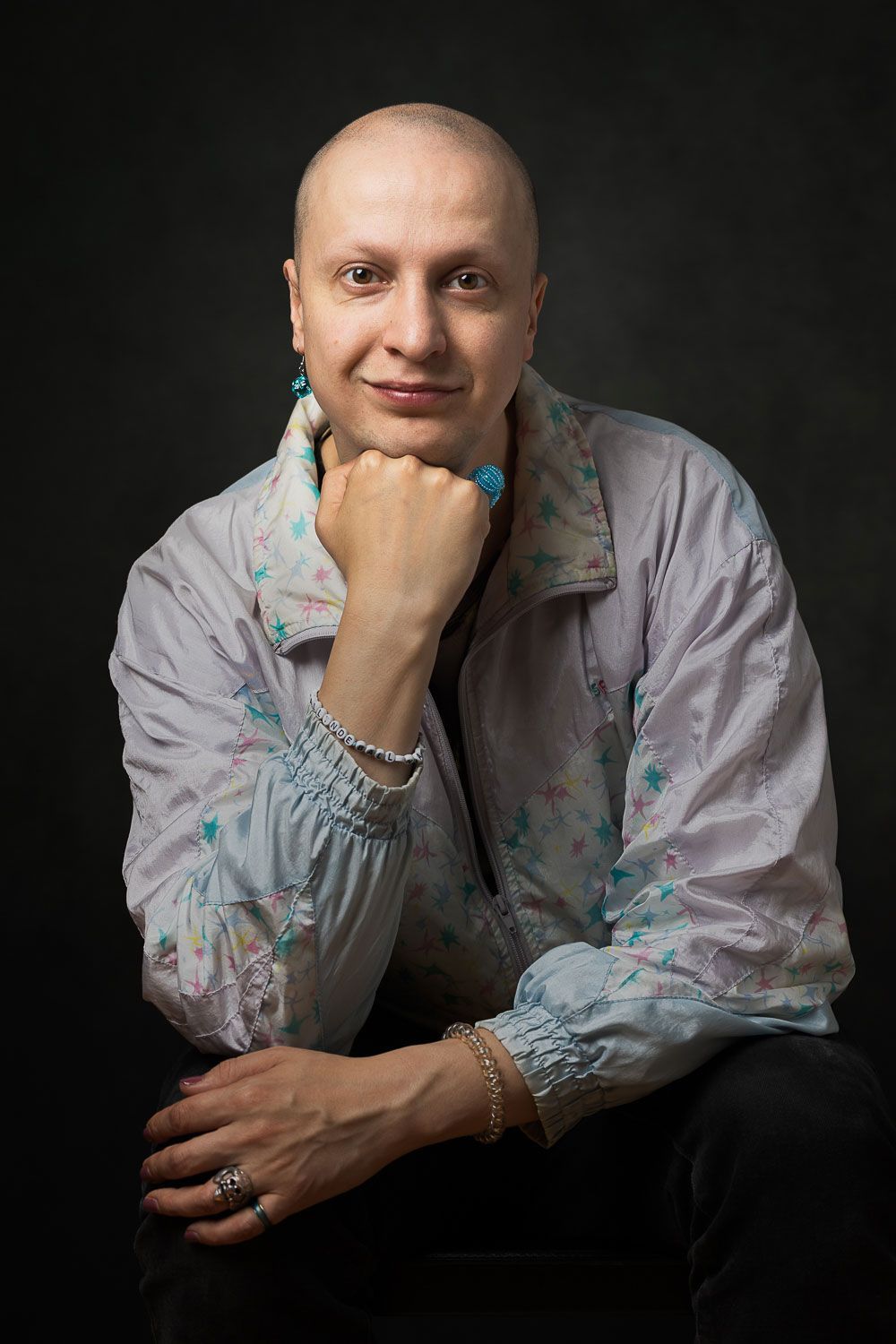 Persoon met geschoren hoofd, gekleed in een lichtgekleurd jasje en sieraden, zijn kin in zijn hand, kijkend naar de toeschouwer.