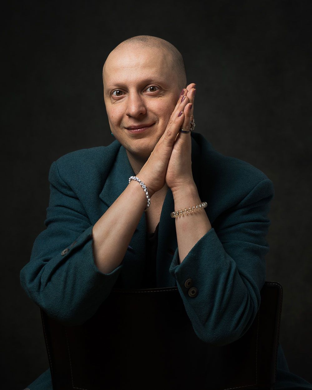 Kaal persoon in een blauwgroen pak, handen gevouwen voor het gezicht, glimlachend. Donkere achtergrond.