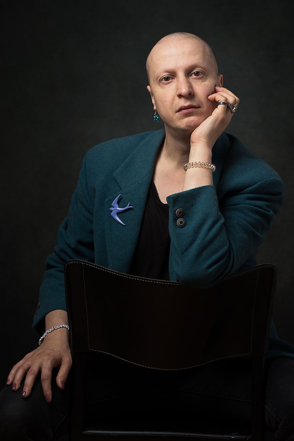 Man met kale kop, blauwgroene jas, hand onder de kin, zittend. Donkere achtergrond.