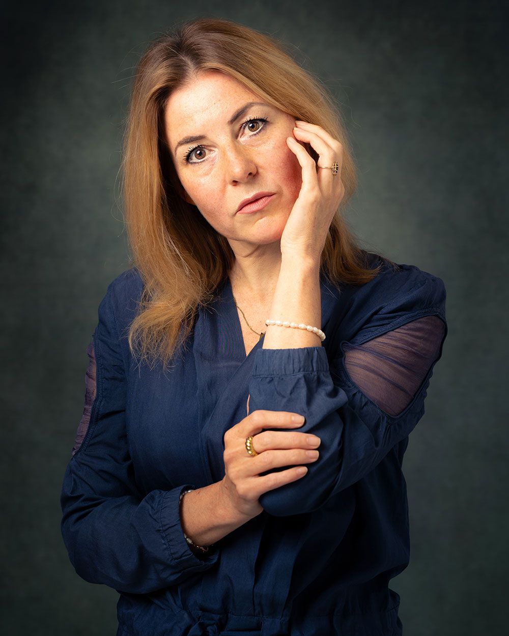 Vrouw in blauw topje, hand op gezicht, kijkend naar de toeschouwer. Donkere achtergrond.