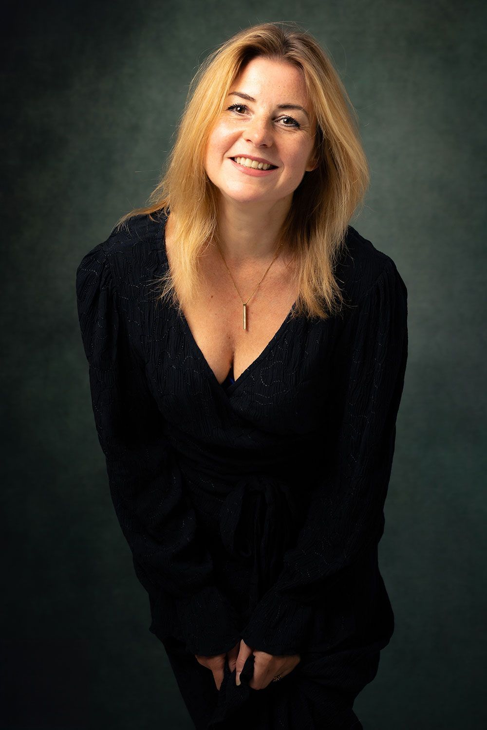 Vrouw met blond haar, glimlachend, gekleed in een donkerblauwe trui, poserend tegen een donkergroene achtergrond.