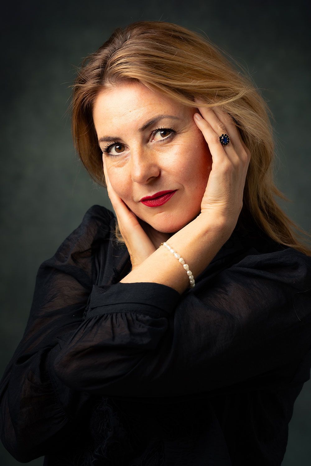 Vrouw met blond haar, handen dicht bij haar gezicht, draagt een zwart shirt en een parelarmband. Donkere achtergrond.