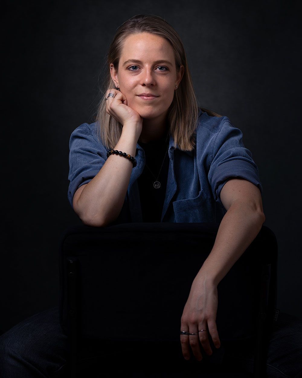 Een vrouw in een spijkerjasje rust met haar kin op haar hand, zittend tegen een donkere achtergrond.