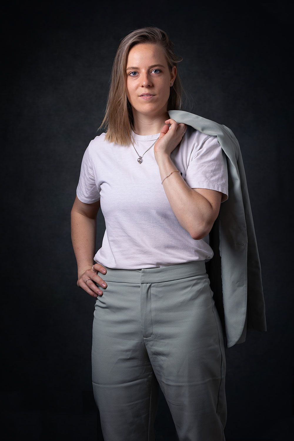Vrouw in een lichte broek en een t-shirt, een blazer over haar schouder en haar hand in haar zij, tegen een donkere achtergrond.