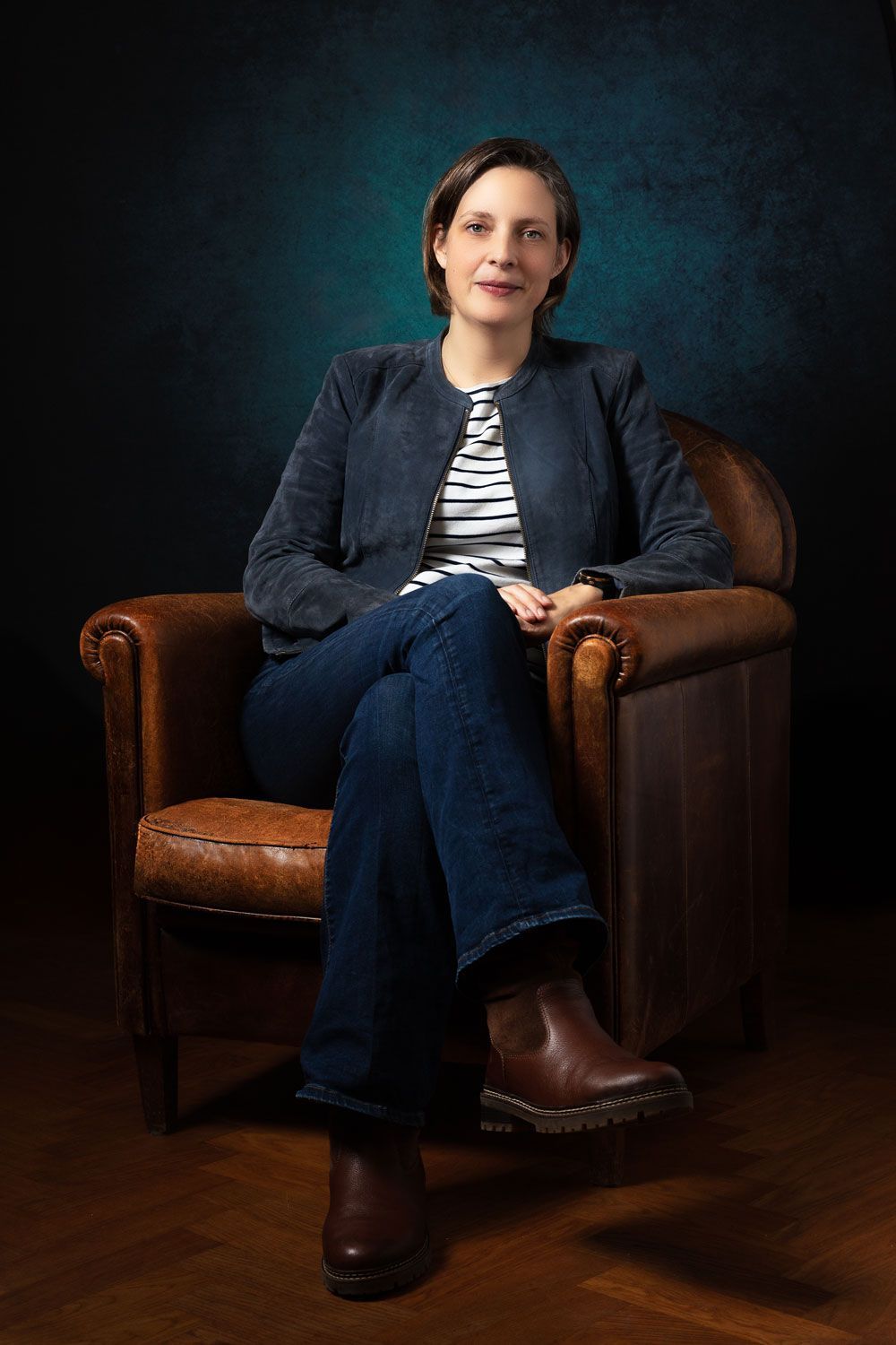 Vrouw zit in een leren fauteuil, gekleed in een blauwe jas, spijkerbroek, gestreept shirt en bruine laarzen.