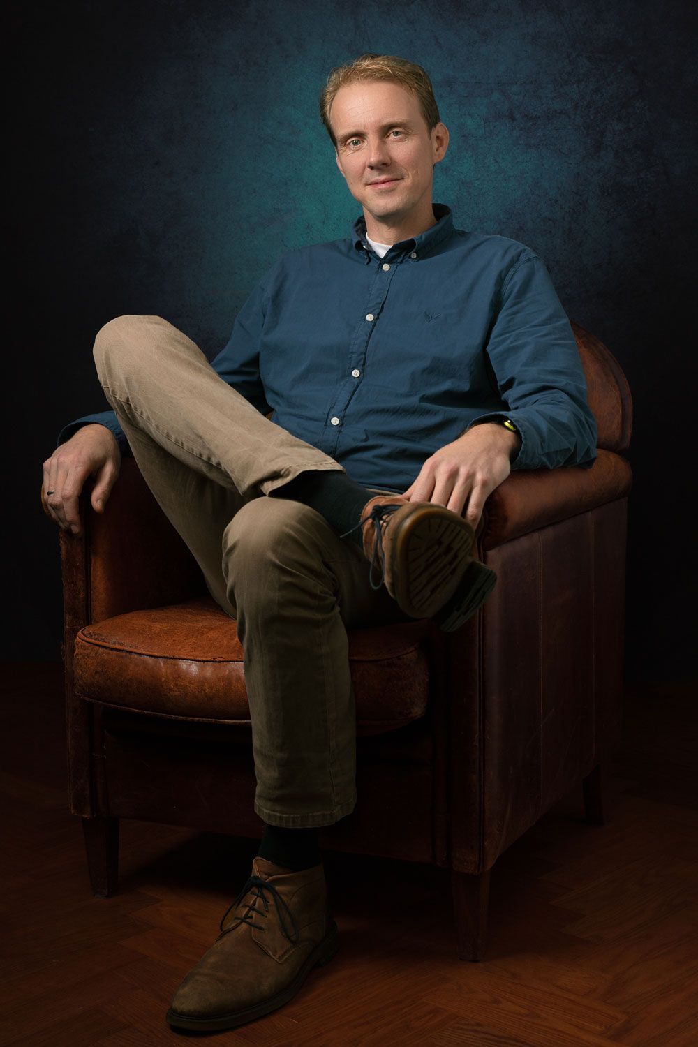 Een man in een blauw shirt en kaki broek zit met gekruiste benen in een leren fauteuil. Donkere, gestructureerde achtergrond.