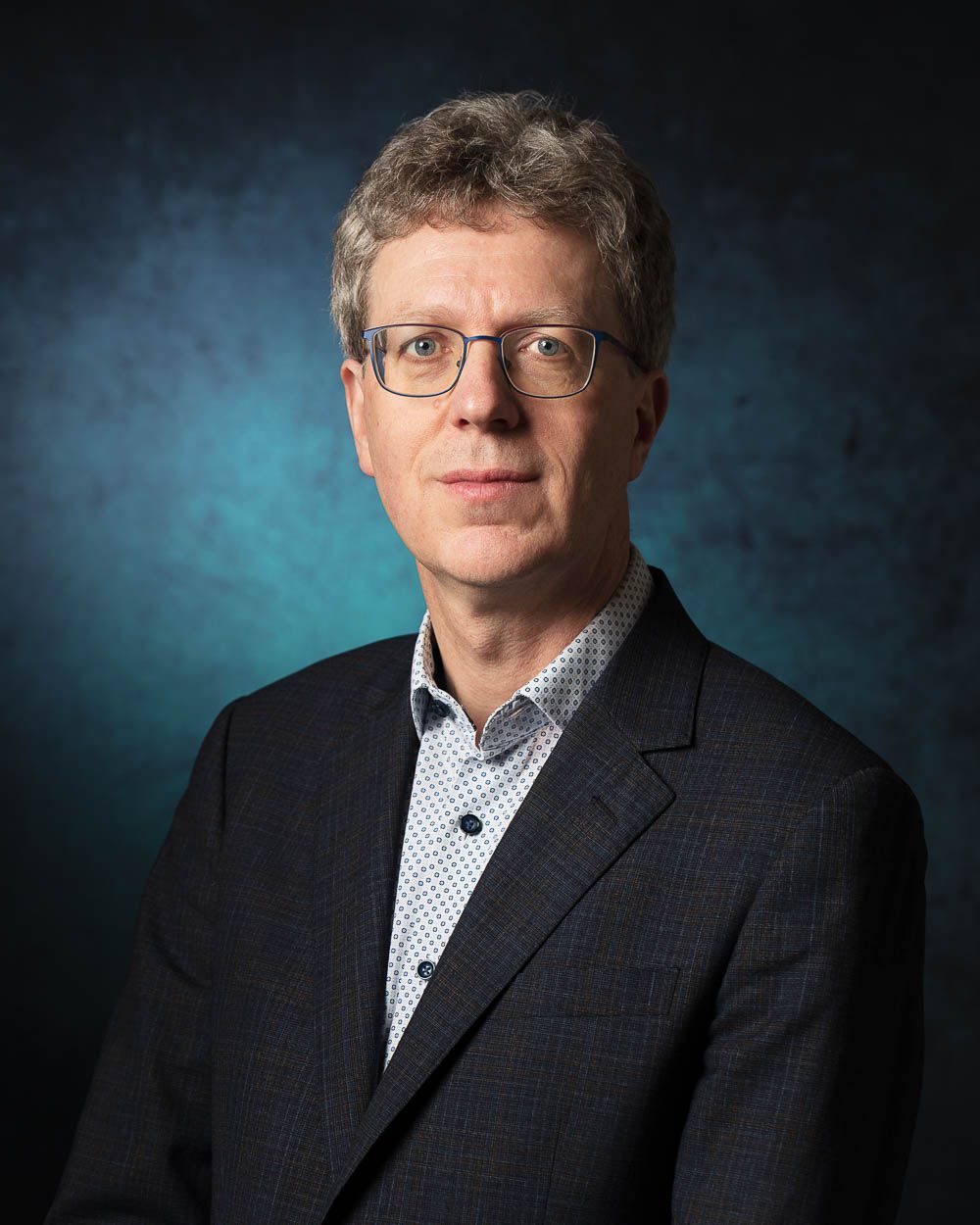 Een man met bril en blazer kijkt in de camera, tegen een donkerblauwe achtergrond.