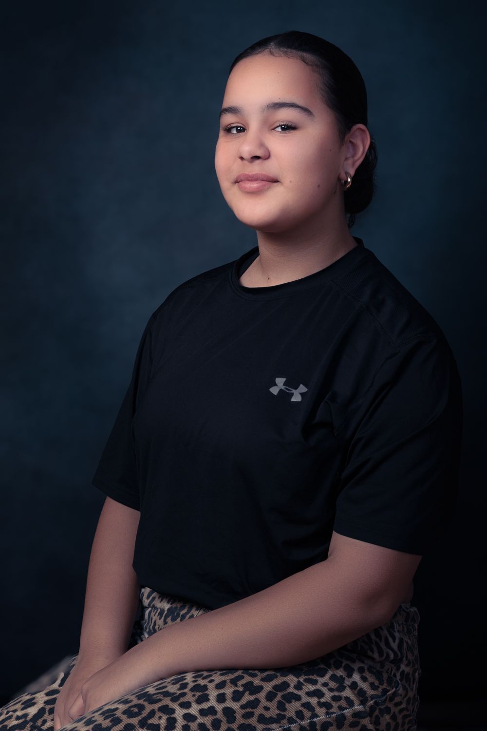 Een zittende persoon, glimlachend, gekleed in een zwart shirt en een rok met luipaardprint, poseert tegen een donkerblauwe achtergrond.