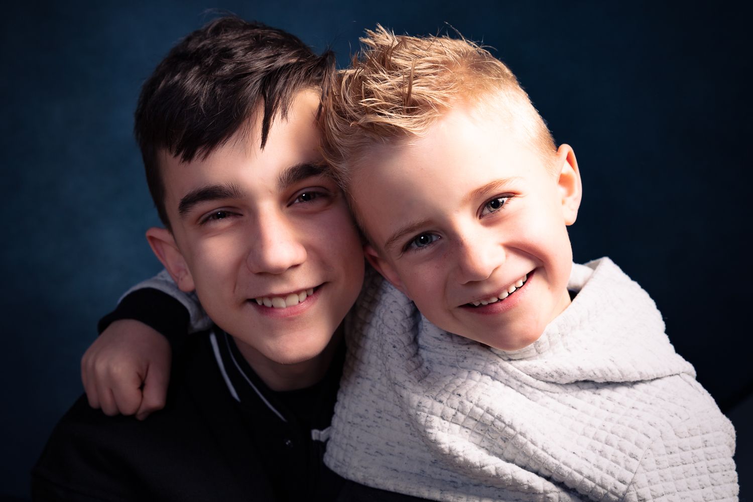 Twee lachende jongens, één met donker haar, met hun armen om de andere jongen met blond haar. Blauwe achtergrond.