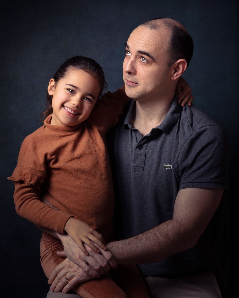 Vader en kind poseren samen. Het kind lacht naar de kijker; de vader kijkt op. Blauwgroene achtergrond.