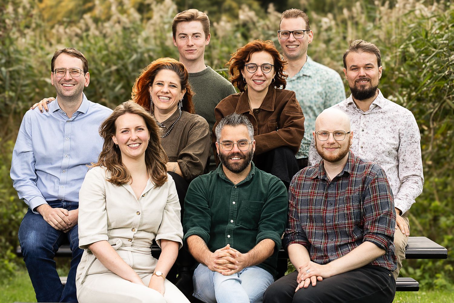 Een groep van tien personen, lachend, poseren buiten voor een groene achtergrond.
