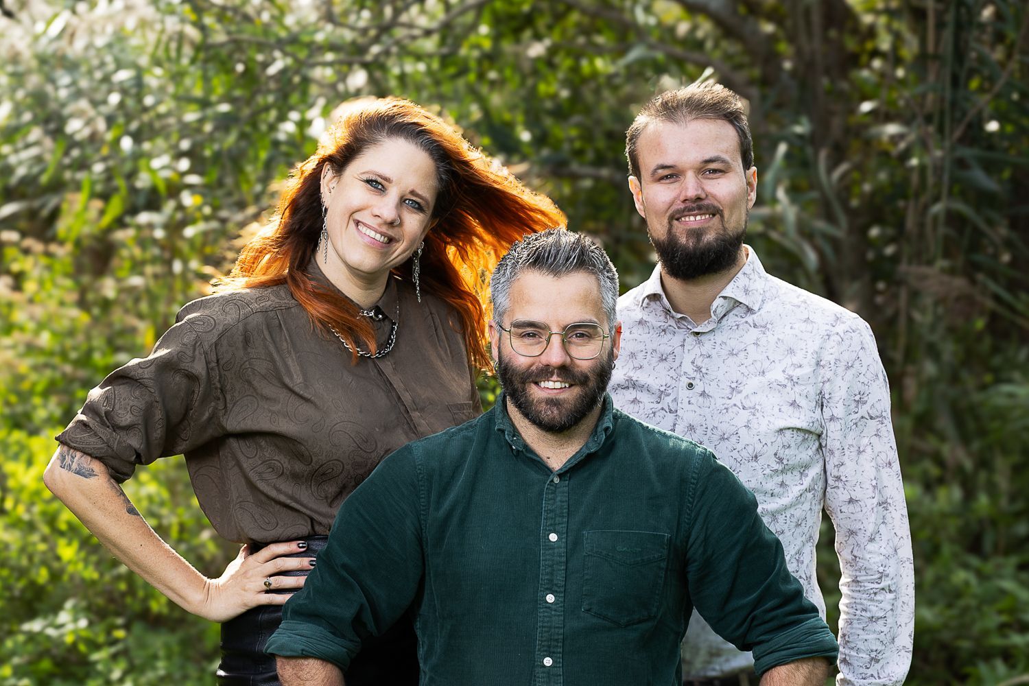 Drie mensen lachen buiten: een vrouw in bruin, een man in groen, een man in een overhemd met een print.
