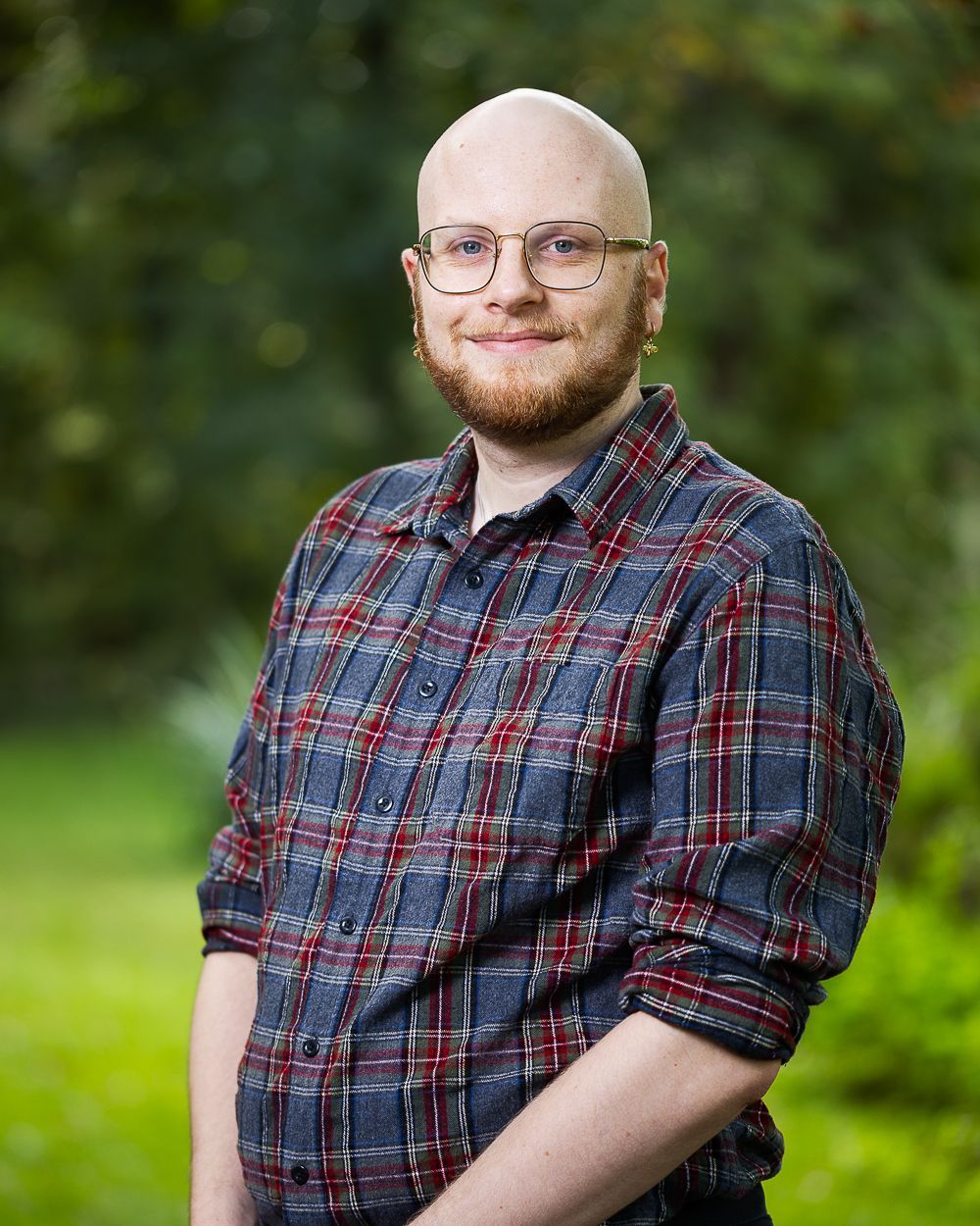 Kale persoon met een baard, een bril en een geruit overhemd, glimlachend buitenshuis.