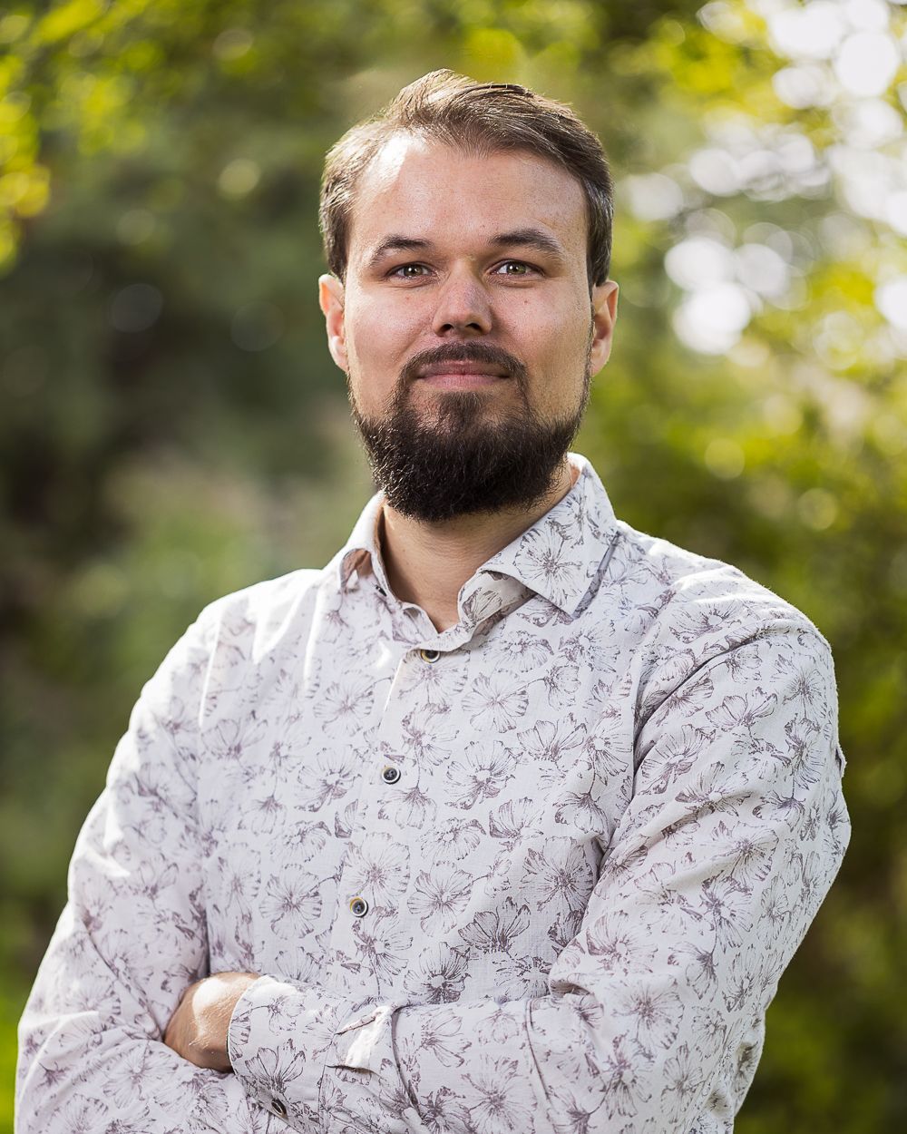 Man met baard in gebloemd overhemd, armen over elkaar, lachend buiten tegen een wazige groene achtergrond.