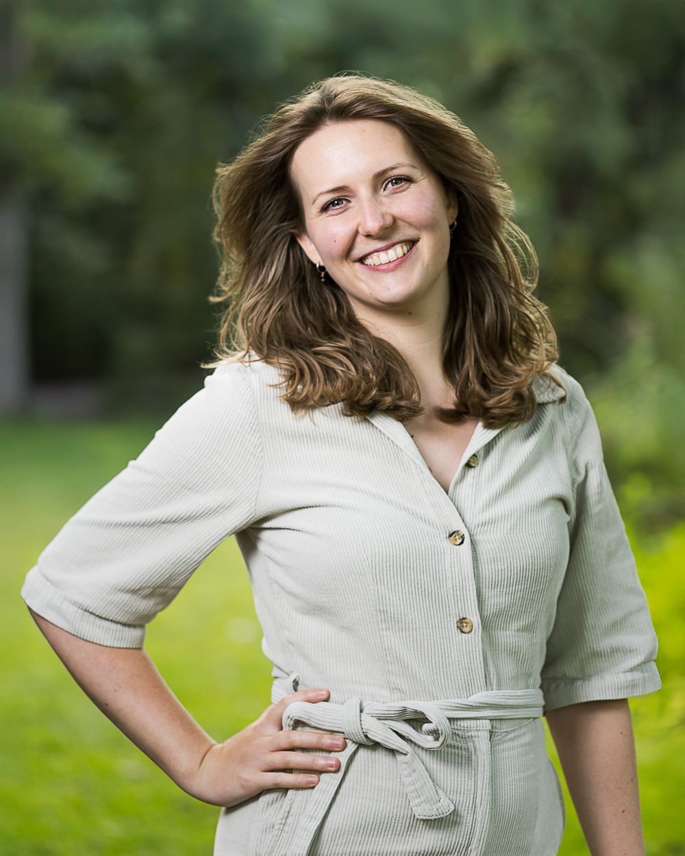 Een vrouw in een lichtgekleurde jurk lacht, haar hand in haar zij, terwijl ze buiten staat voor het groene gebladerte.