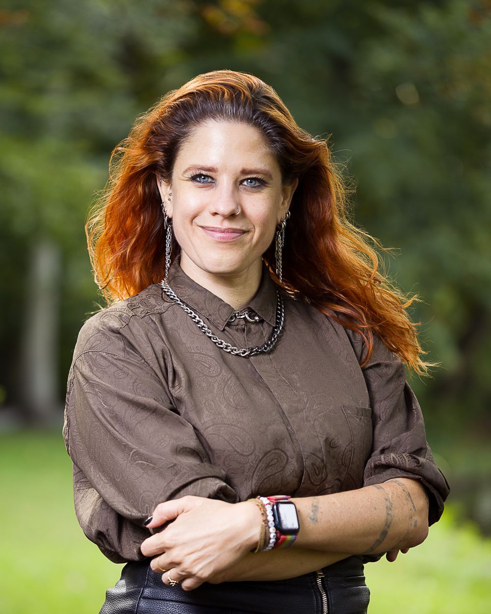 Vrouw met rood haar en bruin shirt voor een wazig groene achtergrond, armen over elkaar, glimlachend.