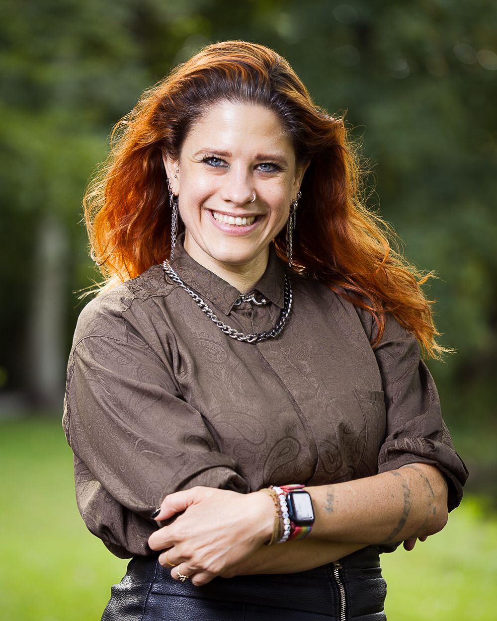 Vrouw met rood haar, glimlachend in de buitenlucht, draagt ​​een bruin shirt en sieraden.