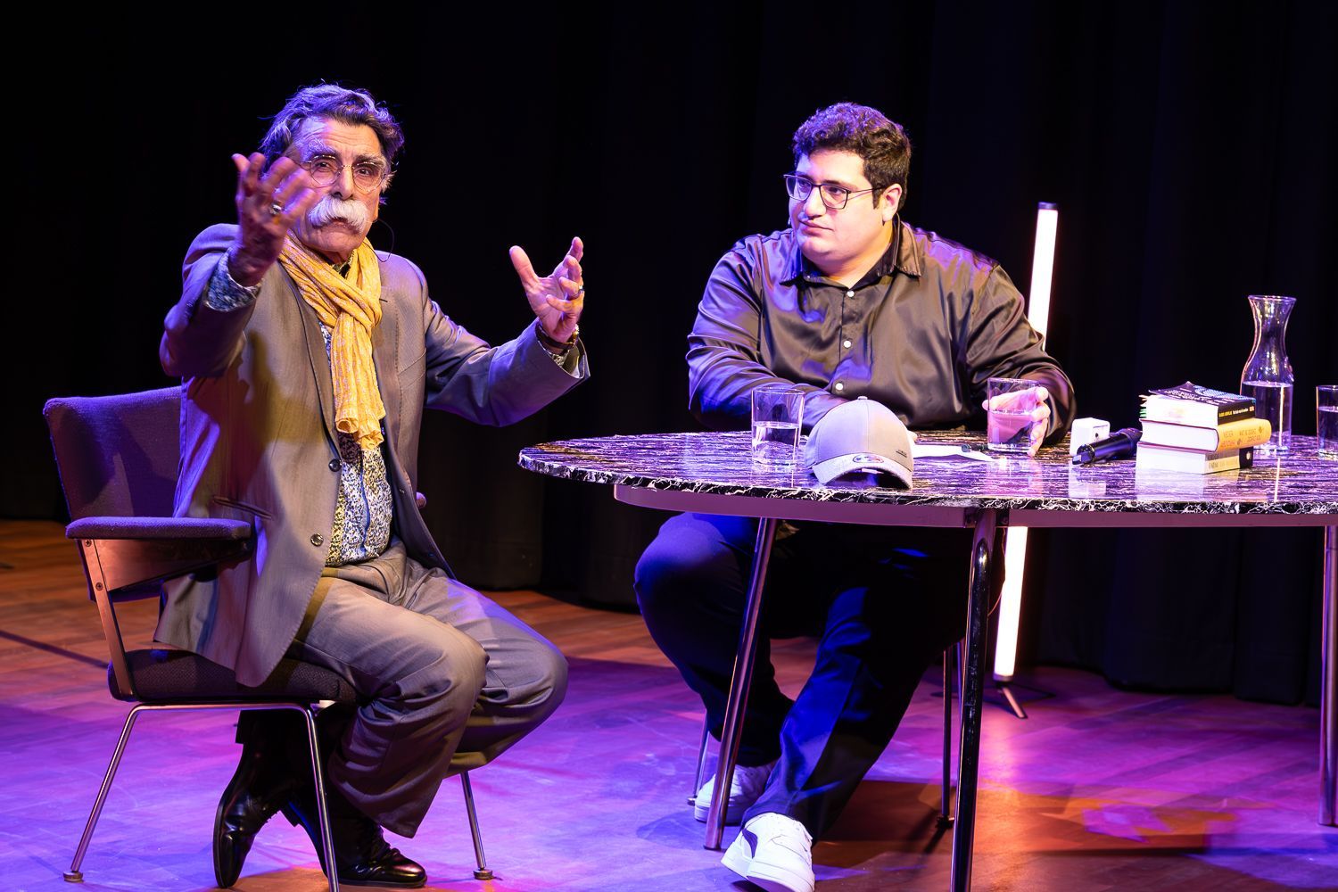Twee mannen zitten aan een tafel op een podium. De een maakt brede gebaren, de ander kijkt toe. Het podium is donker.