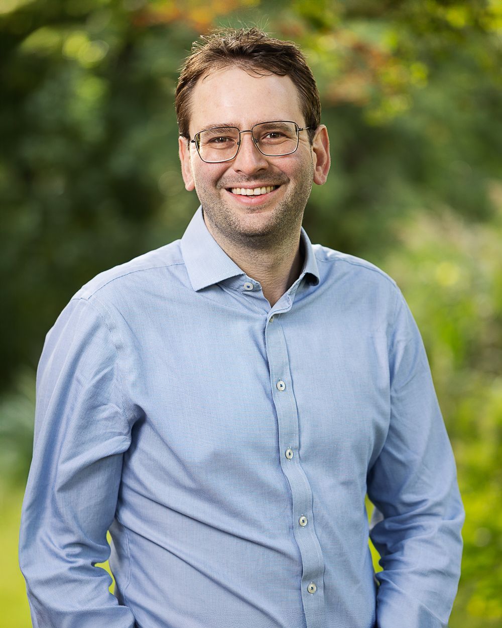 Lachende man met bril, gekleed in een blauw overhemd, buiten met wazig groen gebladerte.
