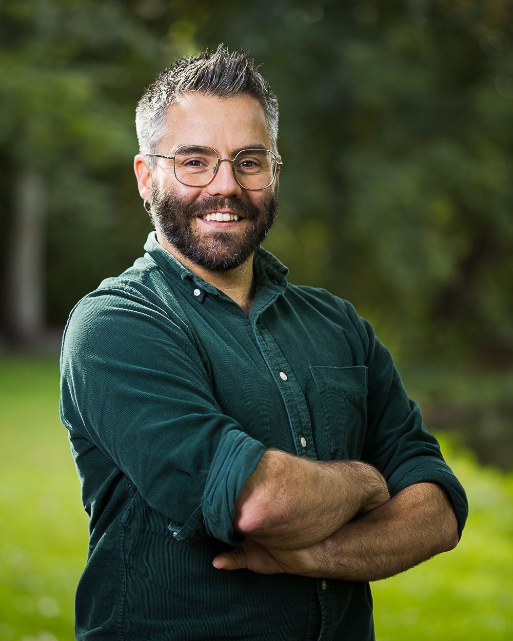 Man met bril en baard, glimlachend, armen over elkaar, gekleed in een groen corduroy overhemd, buiten.