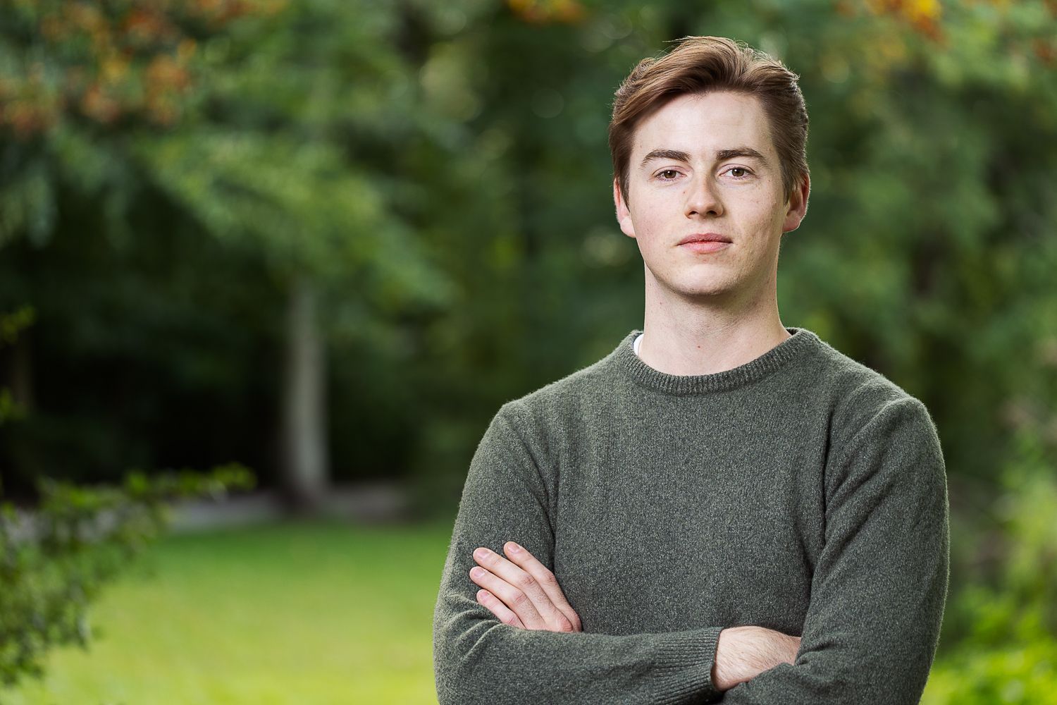 Man met gekruiste armen, gekleed in een grijze trui, staat buiten met bomen op de achtergrond.